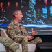 A U.S. Army soldier identified by name tape as 'Ferguson' is seated on a white chair on a stage, actively speaking and gesturing with his hands during what appears to be a panel discussion or conference. He is wearing the Operational Camouflage Pattern (OCP) uniform with a U.S. flag patch and 'U.S. ARMY' tape visible. Another soldier in the same uniform is partially visible on the left side of the frame.