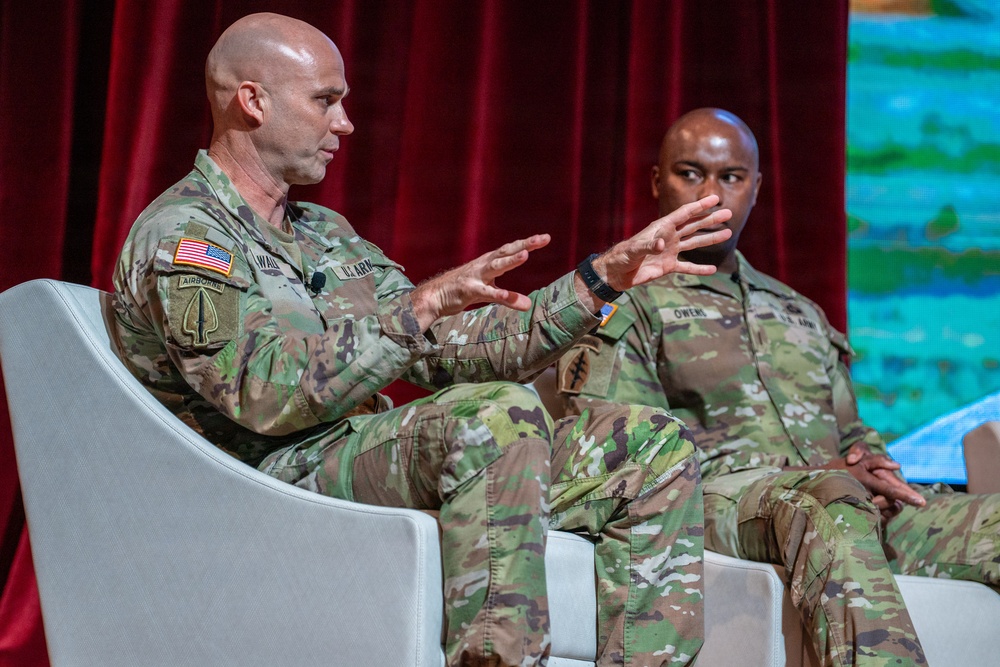 Two U.S. Army soldiers in OCP (Operational Camouflage Pattern) uniforms are seated in white chairs on what appears to be a stage or panel discussion setting. The soldier on the left is actively speaking and gesturing with both hands, while the soldier on the right, whose name tape reads 'OWENS,' listens attentively. The backdrop includes a red curtain and a colorful display screen.