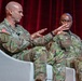 Two U.S. Army soldiers in OCP (Operational Camouflage Pattern) uniforms are seated in white chairs on what appears to be a stage or panel discussion setting. The soldier on the left is actively speaking and gesturing with both hands, while the soldier on the right, whose name tape reads 'OWENS,' listens attentively. The backdrop includes a red curtain and a colorful display screen.