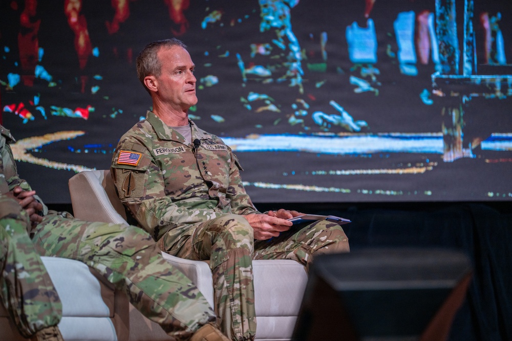 A U.S. Army officer wearing the name tape 'Ferguson' and displaying three stars on his chest (indicating the rank of Lieutenant General) is seated in a white chair on what appears to be a conference or panel stage. He is dressed in OCP (Operational Camouflage Pattern) uniform with a U.S. flag patch on his right shoulder and 'U.S. ARMY' tape visible. A large digital display screen is visible in the background.