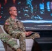 A U.S. Army officer wearing the name tape 'Ferguson' and displaying three stars on his chest (indicating the rank of Lieutenant General) is seated in a white chair on what appears to be a conference or panel stage. He is dressed in OCP (Operational Camouflage Pattern) uniform with a U.S. flag patch on his right shoulder and 'U.S. ARMY' tape visible. A large digital display screen is visible in the background.