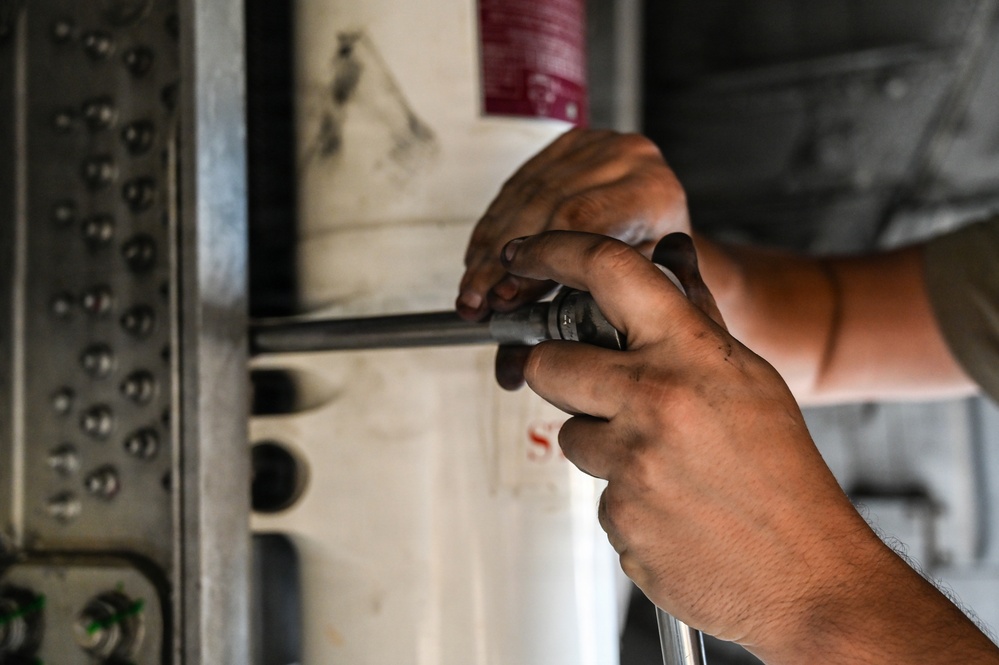 A close-up photograph of a person's hands using a socket wrench or ratchet tool to work on a mechanical or structural component. The subject appears to be performing maintenance or repair work on a large metal structure, possibly an aircraft, vehicle, or industrial equipment. The image is tightly framed, focusing on the hands and tool in action.