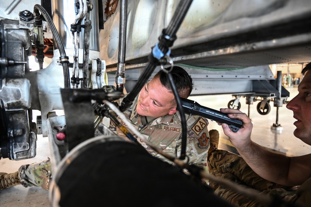 Two U.S. Air Force personnel in OCP (Operational Camouflage Pattern) uniforms are performing maintenance work underneath a military aircraft. One individual, wearing a 'QA' (Quality Assurance) patch, is inspecting components while the other holds what appears to be a flashlight or inspection tool. The work is taking place inside a hangar environment.