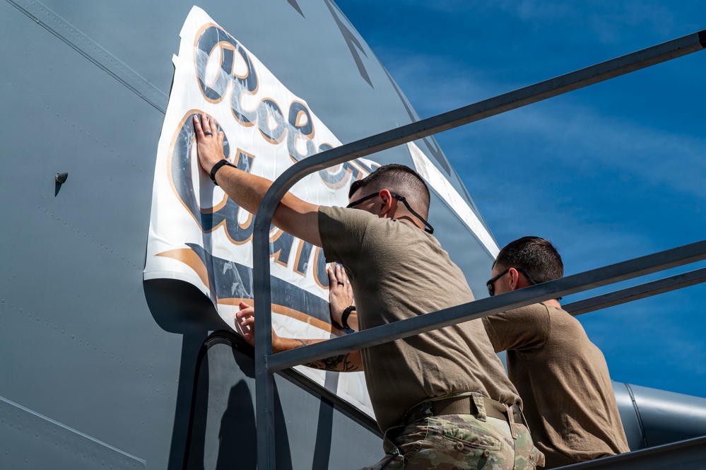 Two individuals in military-style tan/olive uniforms are applying or pressing a large vinyl decal or wrap onto the exterior surface of what appears to be a military aircraft. The decal features circular lettering in blue and gold/bronze colors on a white background. The work is being performed from an elevated platform or maintenance stand outdoors under a blue sky.