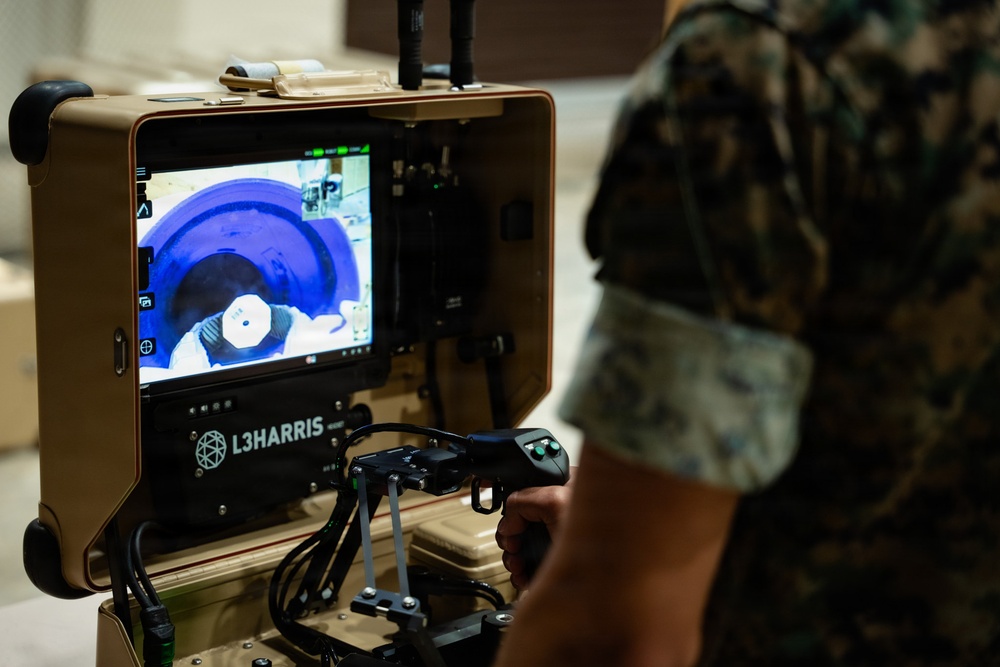 A military personnel in camouflage uniform operates an L3Harris robotic control station, holding a joystick controller while viewing a live feed on the integrated display screen. The screen shows what appears to be a robot's camera perspective examining a circular or cylindrical object, likely in an EOD (Explosive Ordnance Disposal) or reconnaissance context. The equipment is mounted on a tan/desert-colored portable case system.