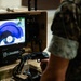 A military personnel in camouflage uniform operates an L3Harris robotic control station, holding a joystick controller while viewing a live feed on the integrated display screen. The screen shows what appears to be a robot's camera perspective examining a circular or cylindrical object, likely in an EOD (Explosive Ordnance Disposal) or reconnaissance context. The equipment is mounted on a tan/desert-colored portable case system.
