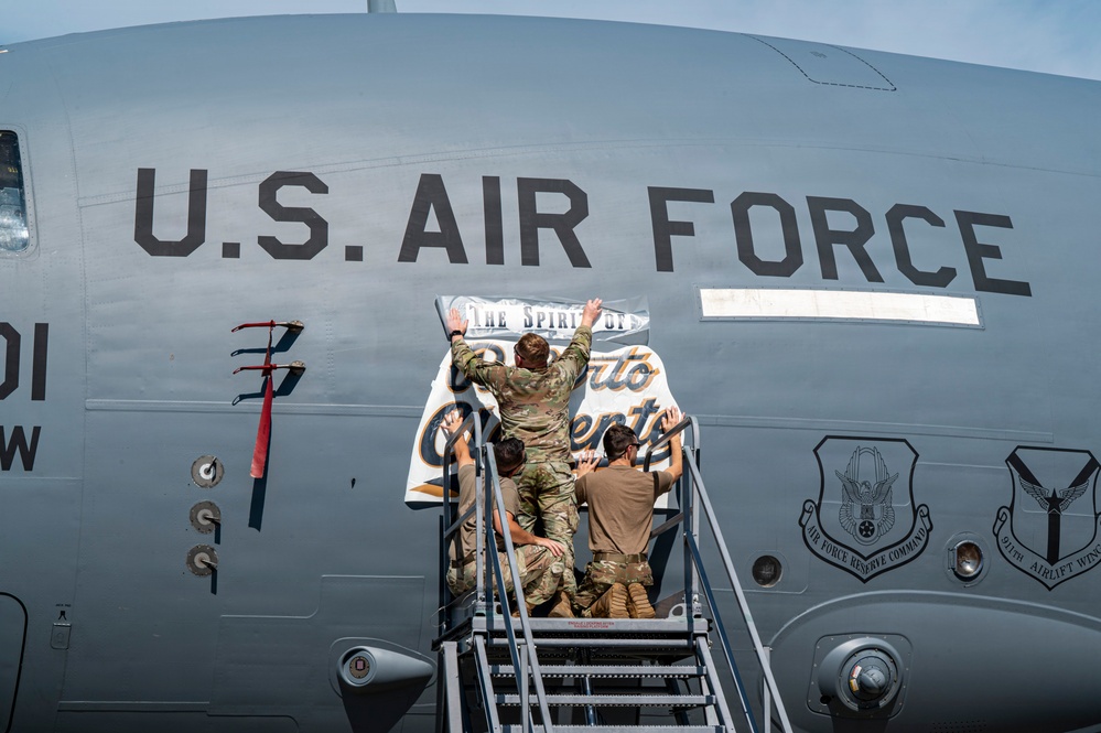 Several U.S. Air Force personnel in military uniforms are standing on a mobile staircase applying or affixing a banner/placard to the fuselage of a large military aircraft. The banner appears to read 'The Spirit of Puerto Rico' or similar text. Two unit insignia patches are visible on the aircraft fuselage, identifying Air Force Reserve Command and the 911th Airlift Wing.