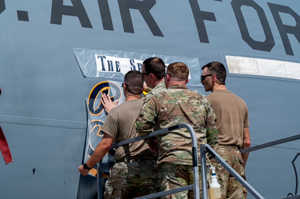 Four military personnel in U.S. Air Force uniforms are applying or adjusting a decal/sticker on the fuselage of a military aircraft. The aircraft bears the text 'U.S. AIR FORCE' and a partially visible name plate reading 'The Sp...' along with a squadron emblem. The personnel are standing on a mobile staircase platform while working on the aircraft exterior.