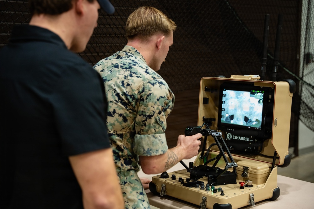 A person in U.S. Marine Corps MARPAT camouflage uniform operates an L3Harris branded portable ground control station, while a second individual in a black polo shirt observes. The ruggedized tan case-style control station features a monitor displaying what appears to be drone or robot camera feed, along with joysticks, buttons, and a wireless antenna. The setting appears to be an indoor demonstration or training environment with chain-link fencing visible in the background.