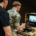 A person in U.S. Marine Corps MARPAT camouflage uniform operates an L3Harris branded portable ground control station, while a second individual in a black polo shirt observes. The ruggedized tan case-style control station features a monitor displaying what appears to be drone or robot camera feed, along with joysticks, buttons, and a wireless antenna. The setting appears to be an indoor demonstration or training environment with chain-link fencing visible in the background.