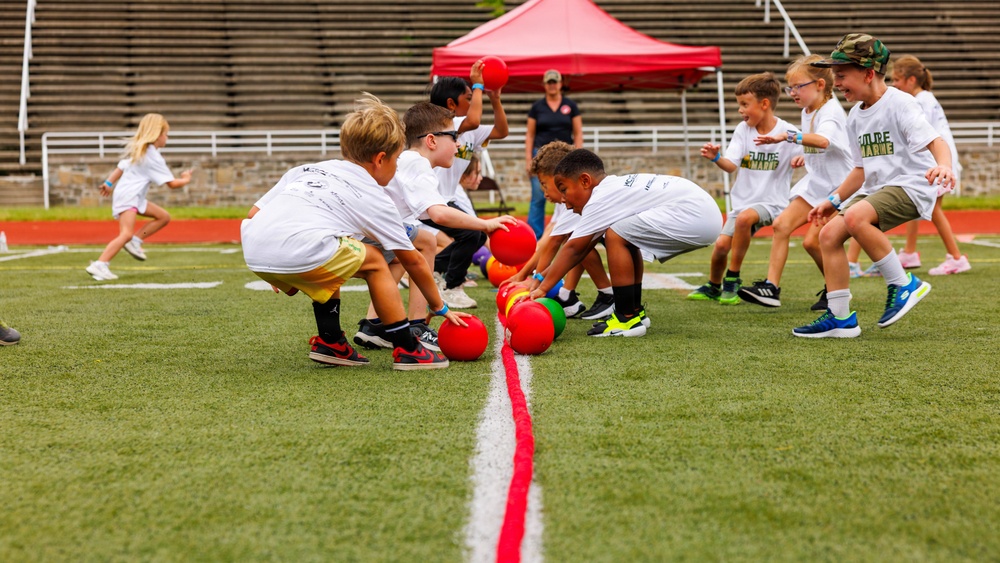 A group of young children are participating in an organized dodgeball game on a green turf athletic field. The children, wearing matching white t-shirts, are scrambling to grab colorful red, orange, and green balls from a center line. An adult supervisor is visible in the background near a red canopy tent.