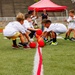 A group of young children are participating in an organized dodgeball game on a green turf athletic field. The children, wearing matching white t-shirts, are scrambling to grab colorful red, orange, and green balls from a center line. An adult supervisor is visible in the background near a red canopy tent.