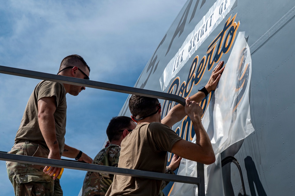 Three military personnel in olive/camouflage uniforms are applying or installing a large vinyl graphic wrap onto the fuselage of an aircraft. The graphic displays the name 'Roberto' in blue and gold script lettering, suggesting a commemorative or tribute marking. The work is being performed from an elevated platform or maintenance stand outdoors under a partly cloudy sky.