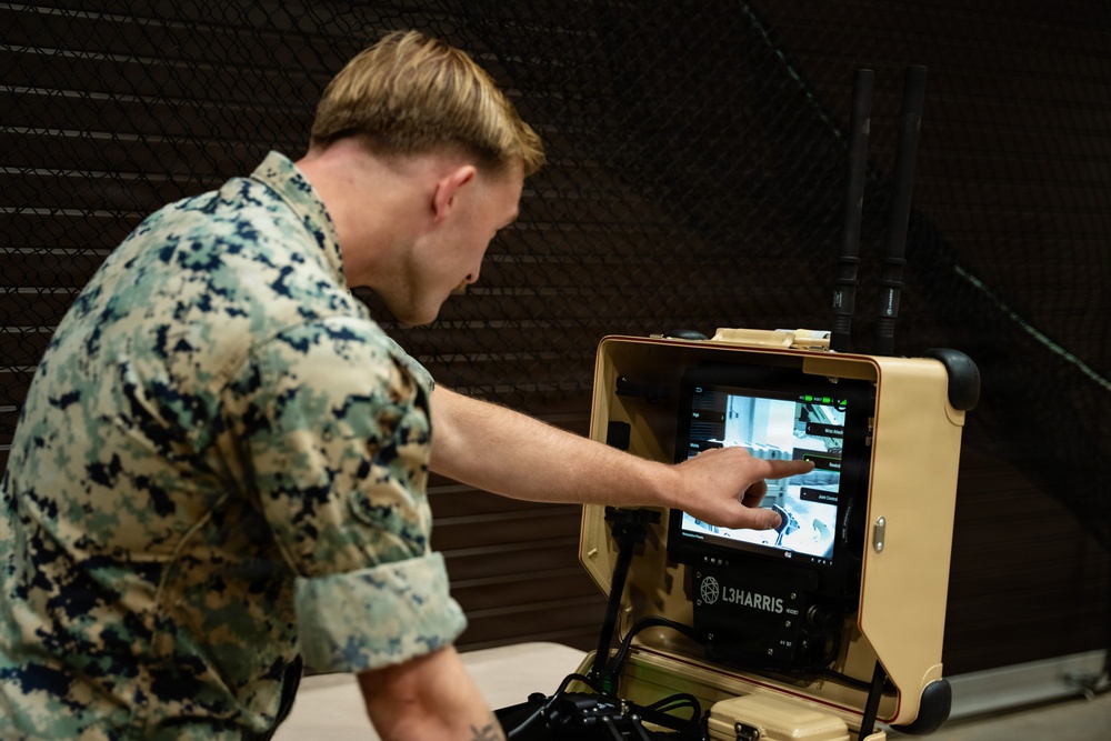 A U.S. Marine in digital camouflage (MARPAT) uniform is operating an L3Harris ruggedized ground control station, touching its tablet display with one finger. The equipment appears to be a portable drone or unmanned system controller housed in a tan/desert-colored hardened case with dual antennas. The setting appears to be an indoor facility with netting visible in the background.