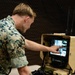 A U.S. Marine in digital camouflage (MARPAT) uniform is operating an L3Harris ruggedized ground control station, touching its tablet display with one finger. The equipment appears to be a portable drone or unmanned system controller housed in a tan/desert-colored hardened case with dual antennas. The setting appears to be an indoor facility with netting visible in the background.