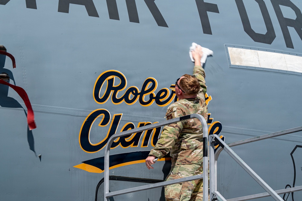 A U.S. Air Force service member in OCP (Operational Camouflage Pattern) uniform is standing on a maintenance platform/ladder, cleaning or polishing the fuselage of a military aircraft. The aircraft bears the name 'Roberto Clemente' in gold and navy blue script lettering, styled in Pittsburgh Pirates baseball team colors. The text 'AIR FORCE' is also visible in large block letters above the name.