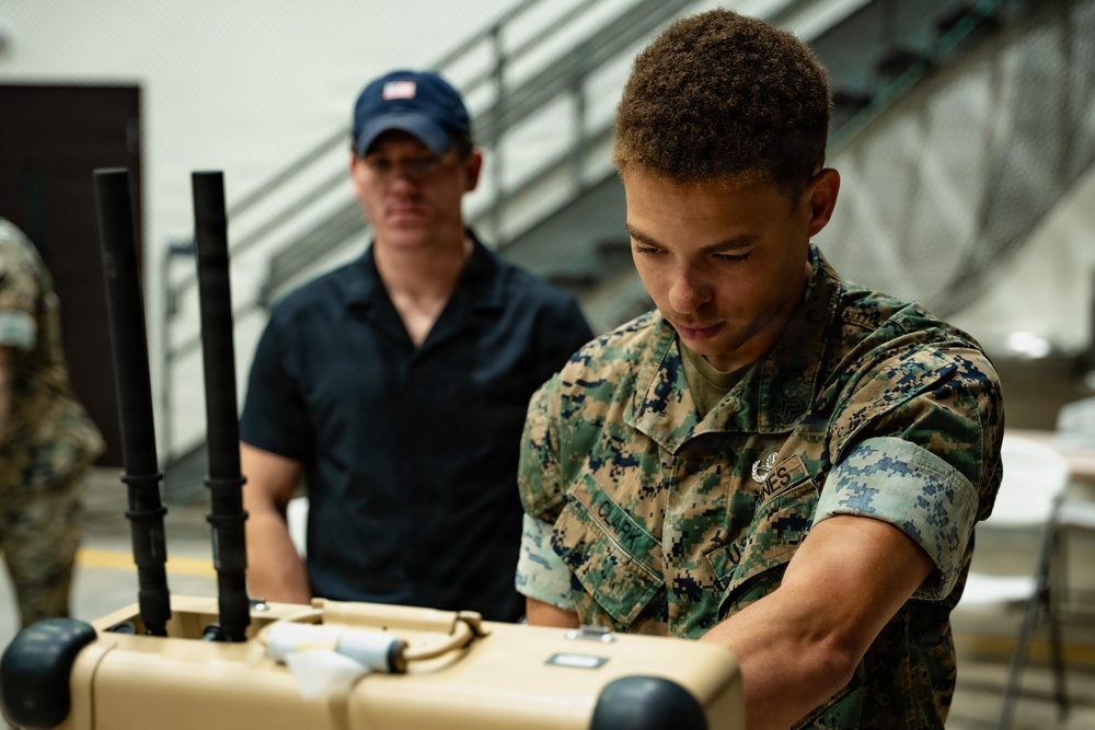A U.S. Marine in MARPAT digital camouflage uniform is handling a tan-colored military equipment case, likely during a setup or inspection activity. A civilian man wearing a dark blue polo shirt and blue baseball cap observes from the background. The setting appears to be an indoor facility with visible staircases and other personnel in the background.