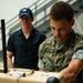 A U.S. Marine in MARPAT digital camouflage uniform is handling a tan-colored military equipment case, likely during a setup or inspection activity. A civilian man wearing a dark blue polo shirt and blue baseball cap observes from the background. The setting appears to be an indoor facility with visible staircases and other personnel in the background.