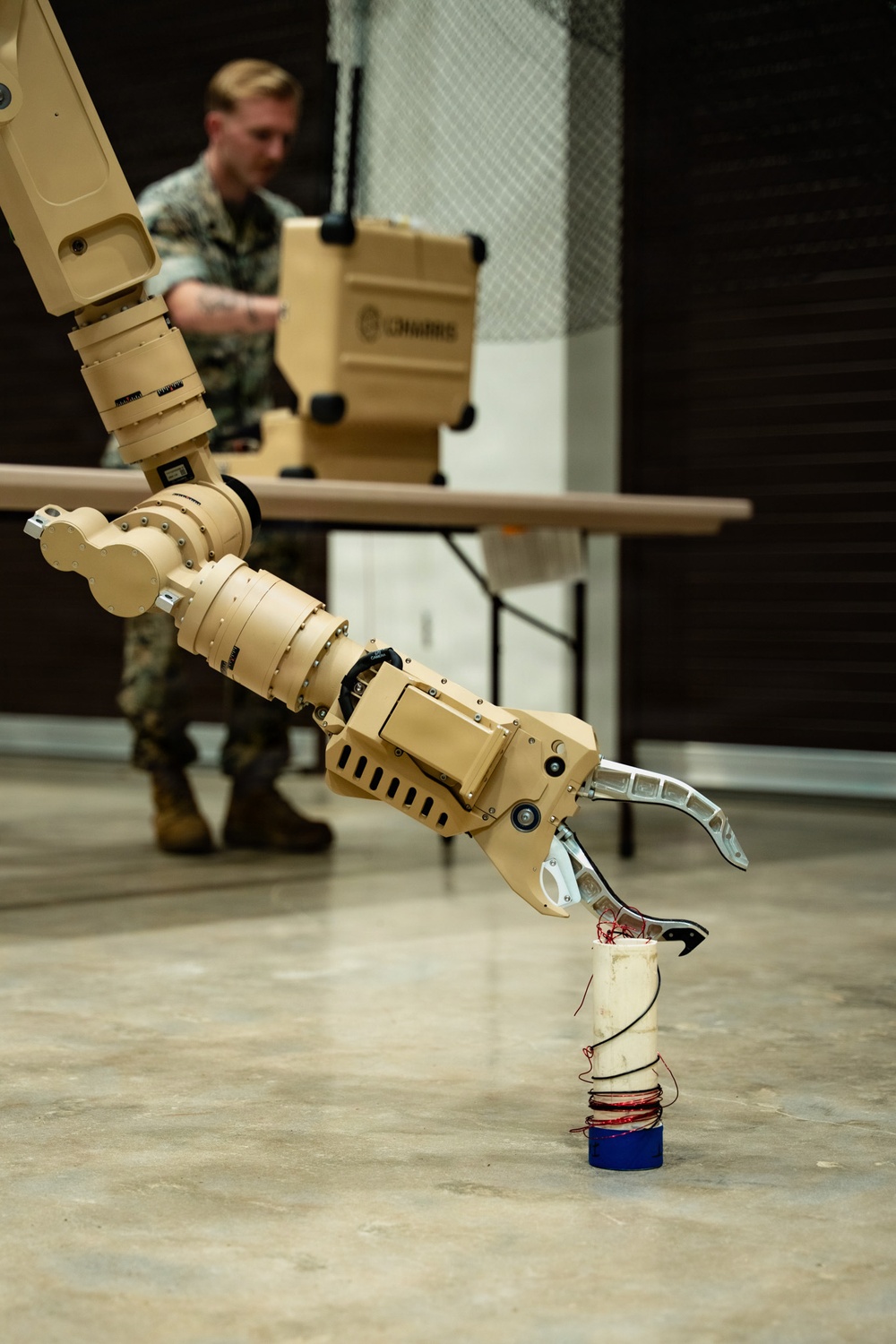 A tan/desert-colored military robotic arm with a claw/gripper end-effector is positioned over a cylindrical object on a concrete floor. The object being manipulated appears to be a white cylinder with red wiring wrapped around it, sitting on a blue base, resembling a simulated IED or training device. In the background, a military service member in camouflage uniform operates what appears to be a control station.