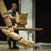 A tan/desert-colored military robotic arm with a claw/gripper end-effector is positioned over a cylindrical object on a concrete floor. The object being manipulated appears to be a white cylinder with red wiring wrapped around it, sitting on a blue base, resembling a simulated IED or training device. In the background, a military service member in camouflage uniform operates what appears to be a control station.