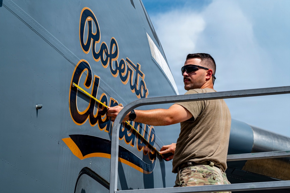 A military service member in camouflage uniform and sunglasses is using a measuring tape on the fuselage of a military aircraft. The aircraft bears the name 'Roberto Clemente' in gold and navy blue script lettering. The photo is taken from a low angle, with the person standing on what appears to be a maintenance platform or stairs.