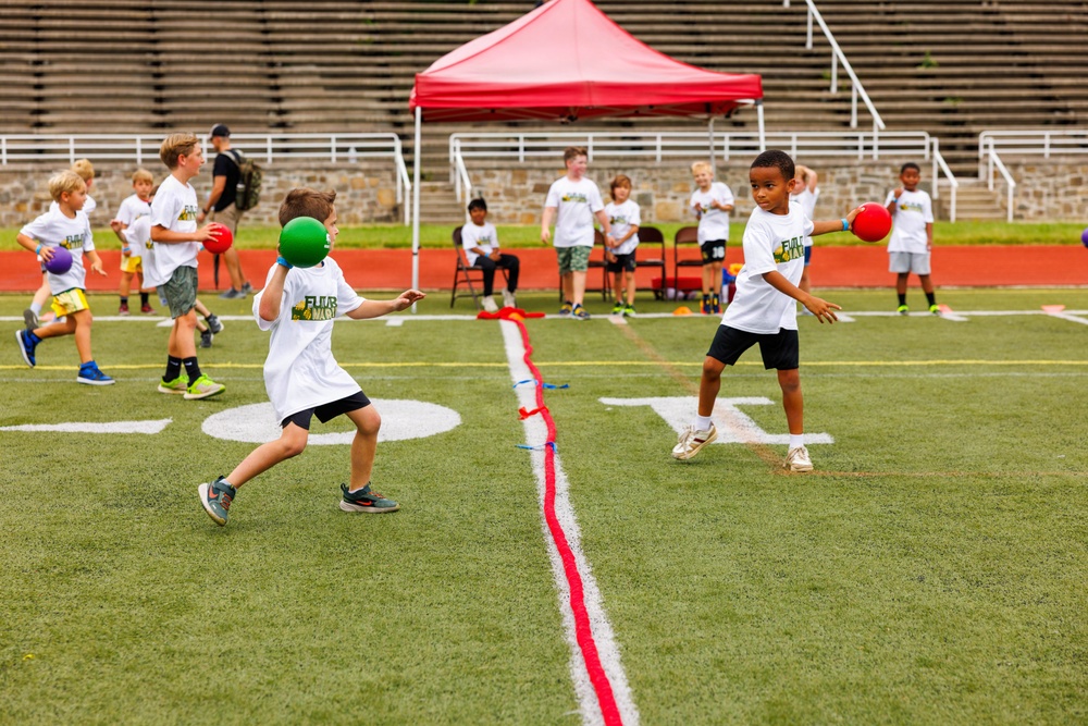 A group of young children are playing dodgeball on a green turf football field, divided by a red center line. All participants are wearing matching white t-shirts with what appears to be a camp or program logo. The event is taking place in a stadium setting with stone bleachers visible in the background, along with a red canopy tent.