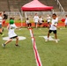 A group of young children are playing dodgeball on a green turf football field, divided by a red center line. All participants are wearing matching white t-shirts with what appears to be a camp or program logo. The event is taking place in a stadium setting with stone bleachers visible in the background, along with a red canopy tent.