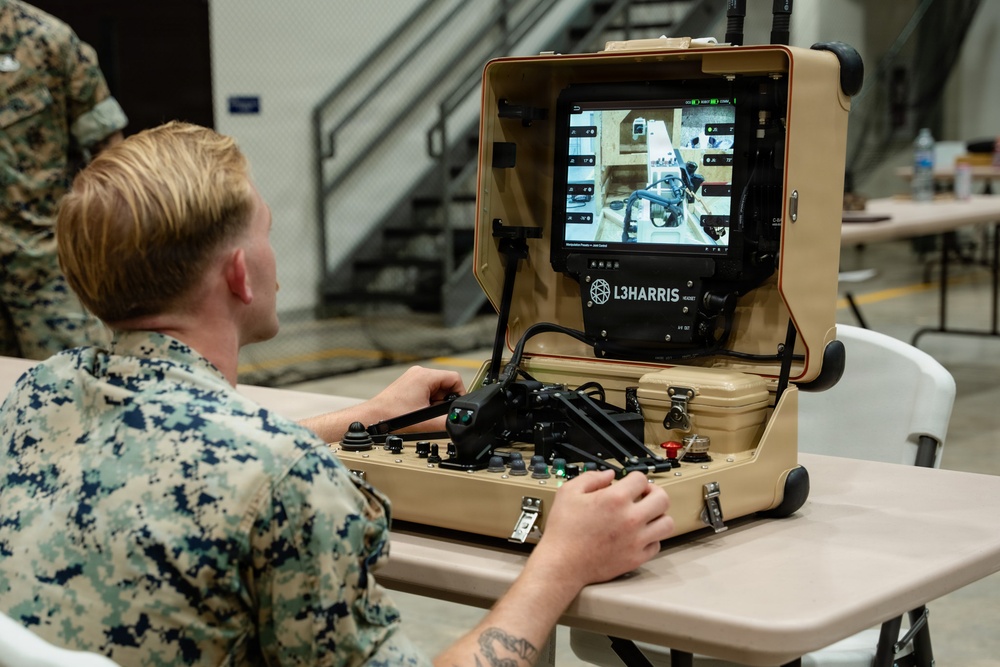 A U.S. Marine in digital camouflage (MARPAT) uniform is operating a portable L3Harris robotic control station at a table in what appears to be an indoor military facility. The suitcase-style control unit features a monitor displaying a live camera feed, joysticks, and various control inputs. The system appears to be used for remote operation of a ground robot or similar unmanned system.
