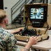 A U.S. Marine in digital camouflage (MARPAT) uniform is operating a portable L3Harris robotic control station at a table in what appears to be an indoor military facility. The suitcase-style control unit features a monitor displaying a live camera feed, joysticks, and various control inputs. The system appears to be used for remote operation of a ground robot or similar unmanned system.