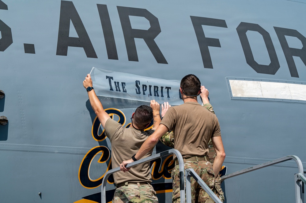 Three U.S. Air Force personnel in tan/multicam uniforms are applying or affixing a decal or banner reading 'The Spirit' onto the fuselage of a U.S. Air Force aircraft. They are standing on a metal staircase/platform to reach the upper portion of the aircraft's body. Additional script lettering in black and gold is visible lower on the fuselage, suggesting a sports team or special livery designation.