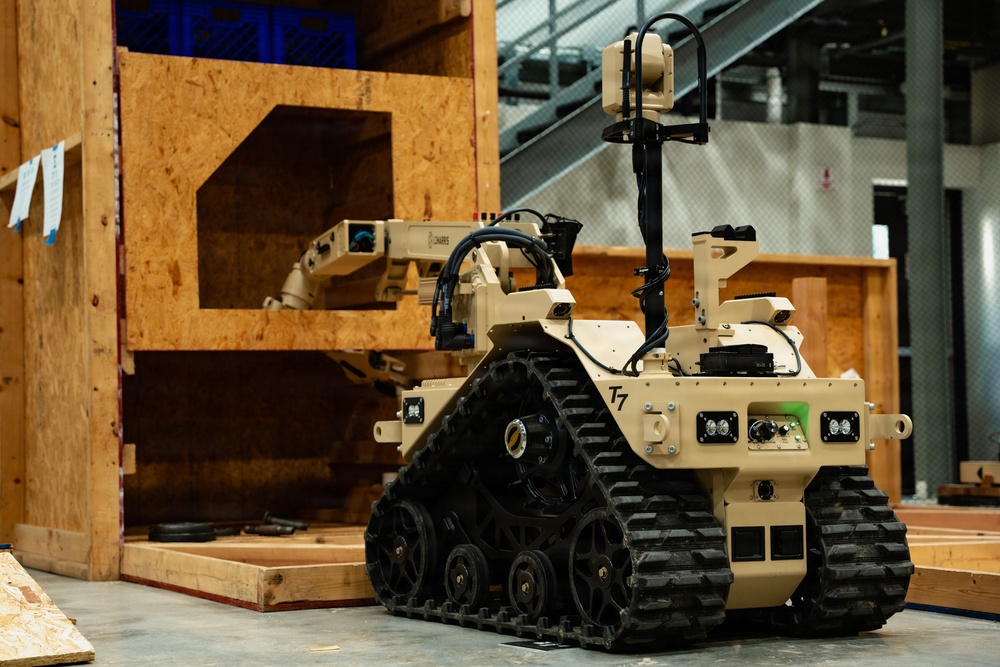 A tan/desert-colored tracked ground robot designated 'T7' is positioned in front of a plywood training structure inside what appears to be an indoor facility. The robot features a robotic arm, multiple cameras, sensors, and rubber tank-style treads. This appears to be a military or law enforcement explosive ordnance disposal (EOD) or reconnaissance robot during a training or demonstration exercise.