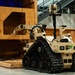 A tan/desert-colored tracked ground robot designated 'T7' is positioned in front of a plywood training structure inside what appears to be an indoor facility. The robot features a robotic arm, multiple cameras, sensors, and rubber tank-style treads. This appears to be a military or law enforcement explosive ordnance disposal (EOD) or reconnaissance robot during a training or demonstration exercise.
