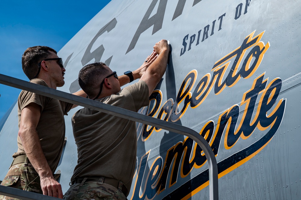 Two U.S. Air Force personnel in olive/tan military uniforms are applying or smoothing a decorative decal onto the fuselage of a military aircraft. The aircraft bears the text 'Spirit of Roberto Clemente' in gold and navy blue script lettering, honoring the legendary baseball player. The work is being performed on a raised platform or stairs alongside the aircraft under clear blue skies.