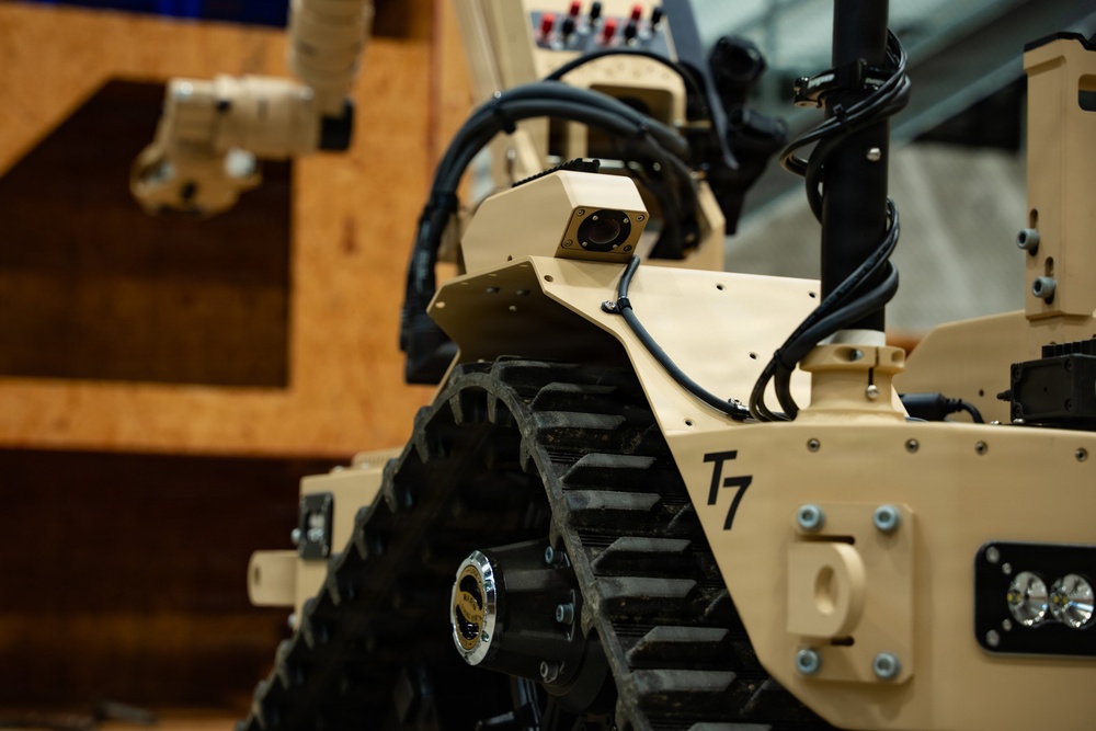 The image shows a close-up view of a military/tactical ground robot designated 'T7', featuring a tan/desert-colored chassis with rubber track drive system. The robot is equipped with a camera module, cable harnesses, LED lighting, and what appears to be a robotic arm visible in the background. The setting appears to be an indoor exhibition or demonstration environment.