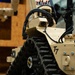 The image shows a close-up view of a military/tactical ground robot designated 'T7', featuring a tan/desert-colored chassis with rubber track drive system. The robot is equipped with a camera module, cable harnesses, LED lighting, and what appears to be a robotic arm visible in the background. The setting appears to be an indoor exhibition or demonstration environment.