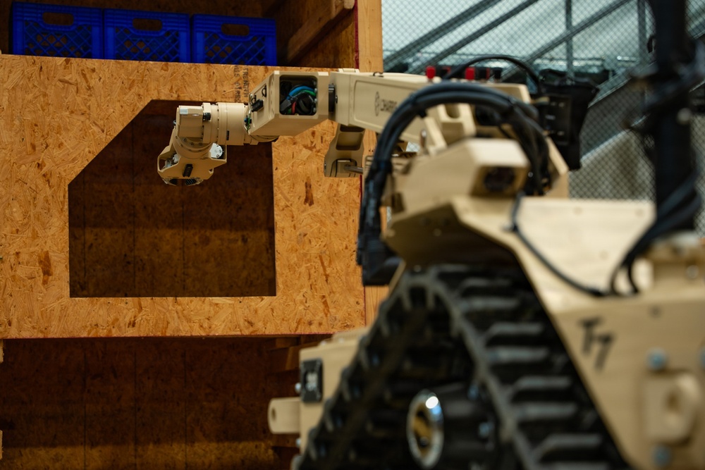 A tan/desert-colored military or tactical ground robot with a tracked chassis is extending a multi-jointed robotic arm through a rectangular cutout in an OSB (oriented strand board) plywood wall. The robot appears to be performing a manipulation or reconnaissance task, likely in a training or testing environment. Blue plastic crates are visible on a shelf in the background.