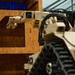 A tan/desert-colored military or tactical ground robot with a tracked chassis is extending a multi-jointed robotic arm through a rectangular cutout in an OSB (oriented strand board) plywood wall. The robot appears to be performing a manipulation or reconnaissance task, likely in a training or testing environment. Blue plastic crates are visible on a shelf in the background.