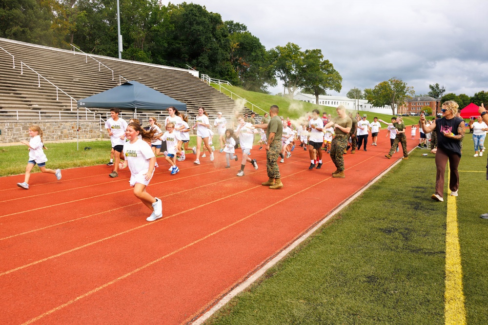 A group of children in white t-shirts are running on a red athletic track, participating in what appears to be a color run or fun run event. Military personnel in camouflage uniforms are stationed along the track throwing colored powder at the runners. The event appears to take place at a military installation, evidenced by the stadium bleachers and institutional buildings visible in the background.
