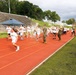 A group of children in white t-shirts are running on a red athletic track, participating in what appears to be a color run or fun run event. Military personnel in camouflage uniforms are stationed along the track throwing colored powder at the runners. The event appears to take place at a military installation, evidenced by the stadium bleachers and institutional buildings visible in the background.