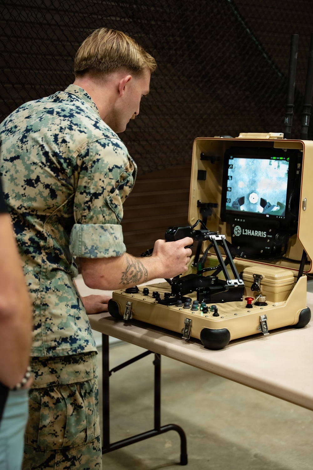 A U.S. Marine in MARPAT digital camouflage uniform is operating an L3Harris ground control station, likely for an unmanned ground vehicle (UGV) or robotic system. The portable tan-colored briefcase-style control unit features joysticks, buttons, and a monitor displaying a live camera feed. The setting appears to be an indoor training or demonstration environment.