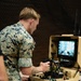 A U.S. Marine in MARPAT digital camouflage uniform is operating an L3Harris ground control station, likely for an unmanned ground vehicle (UGV) or robotic system. The portable tan-colored briefcase-style control unit features joysticks, buttons, and a monitor displaying a live camera feed. The setting appears to be an indoor training or demonstration environment.