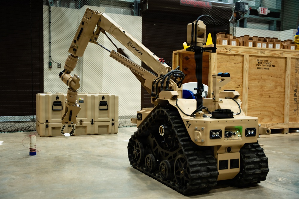 The image shows a tan/desert-colored military ground robot designated 'T7', manufactured by L3Harris, equipped with a multi-jointed robotic arm. The robot is mounted on rubber tank-style tracks and appears to be in an indoor facility, likely a demonstration or testing environment. The robotic arm is extended toward what appears to be a tan military-style equipment case on the floor.