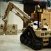 The image shows a tan/desert-colored military ground robot designated 'T7', manufactured by L3Harris, equipped with a multi-jointed robotic arm. The robot is mounted on rubber tank-style tracks and appears to be in an indoor facility, likely a demonstration or testing environment. The robotic arm is extended toward what appears to be a tan military-style equipment case on the floor.