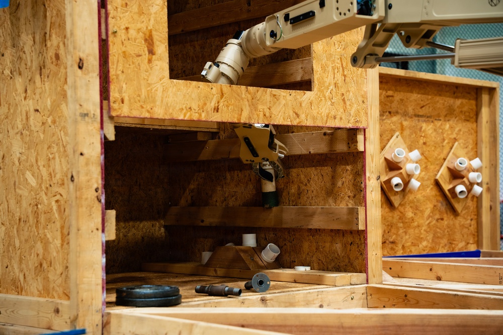 The image shows a robotic arm operating within a wooden OSB (oriented strand board) enclosure or test environment. The robot appears to be performing a manipulation or assembly task involving PVC pipe fittings and connectors. The setup resembles a controlled testing scenario, possibly for construction automation or robotics research.