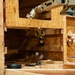 The image shows a robotic arm operating within a wooden OSB (oriented strand board) enclosure or test environment. The robot appears to be performing a manipulation or assembly task involving PVC pipe fittings and connectors. The setup resembles a controlled testing scenario, possibly for construction automation or robotics research.