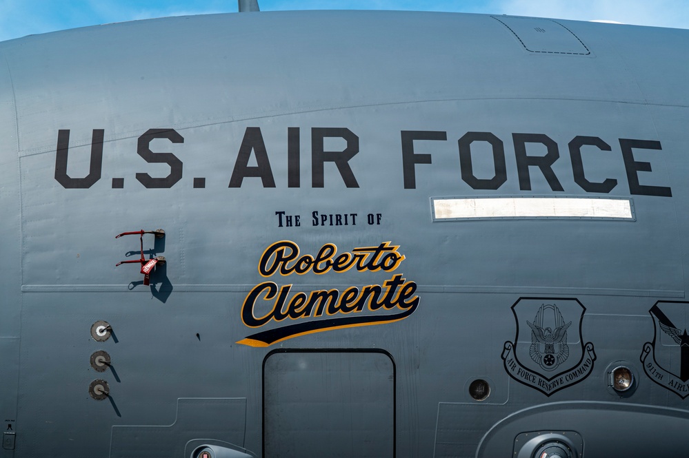 The image shows the fuselage of a U.S. Air Force aircraft named 'The Spirit of Roberto Clemente,' rendered in a baseball-style script with gold and black lettering. The aircraft bears markings from the Air Force Reserve Command, likely the 911th Airlift Wing. The naming honors the legendary Pittsburgh Pirates baseball player and humanitarian Roberto Clemente.