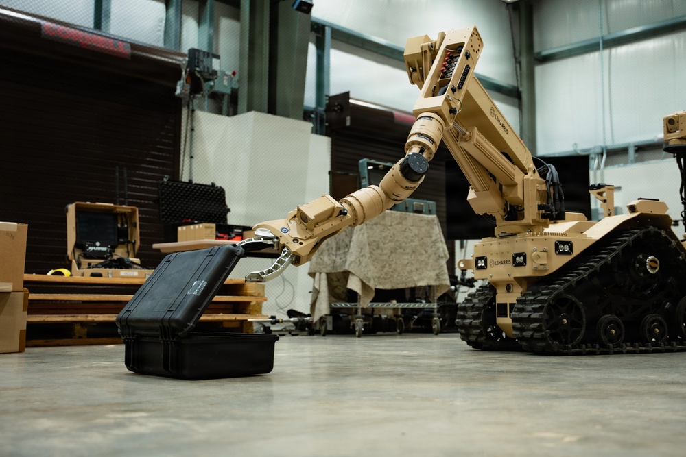 A tan/desert-colored tracked military robot manufactured by L3Harris is shown manipulating a black hard-shell case with its articulated robotic arm in what appears to be an indoor warehouse or demonstration facility. The robot is equipped with rubber tracks for mobility and a multi-jointed arm with a claw/gripper end effector. A second similar robot is partially visible on the right side of the frame.