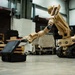 A tan/desert-colored tracked military robot manufactured by L3Harris is shown manipulating a black hard-shell case with its articulated robotic arm in what appears to be an indoor warehouse or demonstration facility. The robot is equipped with rubber tracks for mobility and a multi-jointed arm with a claw/gripper end effector. A second similar robot is partially visible on the right side of the frame.