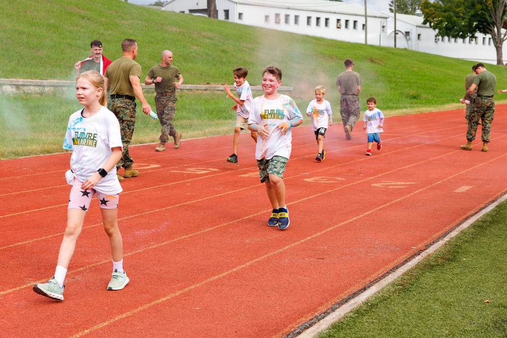 Children wearing 'Future Marine' t-shirts run on a red athletic track alongside adults in military camouflage uniforms. Colored powder is visible in the air, suggesting a color run or fun run event on what appears to be a military base. The children appear joyful and energetic, participating in what looks like a family-friendly military community event.