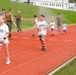 Children wearing 'Future Marine' t-shirts run on a red athletic track alongside adults in military camouflage uniforms. Colored powder is visible in the air, suggesting a color run or fun run event on what appears to be a military base. The children appear joyful and energetic, participating in what looks like a family-friendly military community event.